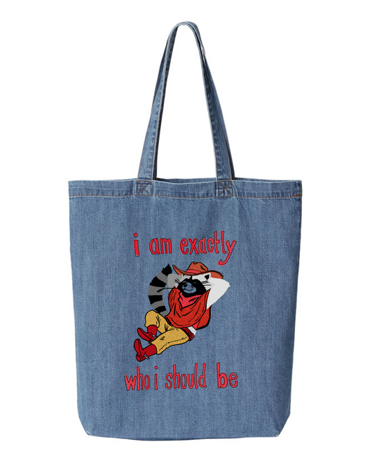 Denim tote bag printed with a raccoon in a cowboy hat and red boots leaning back, with the words i am exactly who i should be
