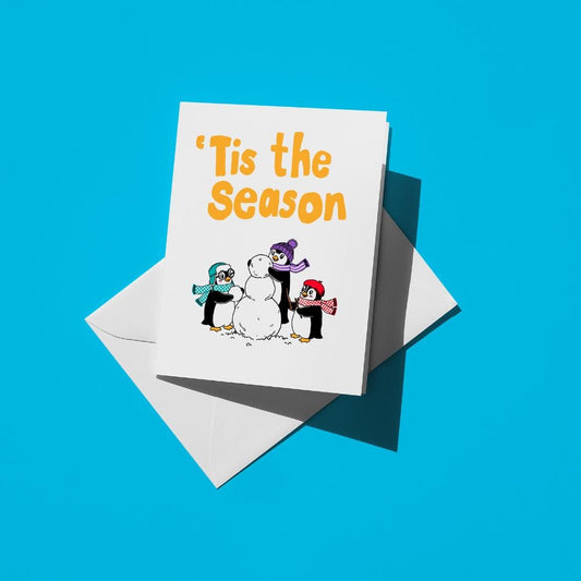 Penguins in scarves building a snowman, with orange text reading "Tis the season"