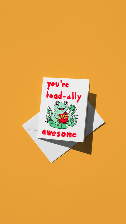 Hand-drawn illustration of a frog on a lilypad holding a box of chocolates with the message "you're toad-ally awesome"